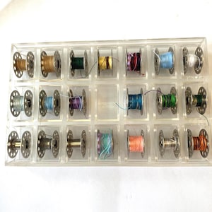 May include: A clear plastic storage case containing seventeen metal sewing machine bobbins, each wound with thread in various colors including gold, green, yellow, purple, blue, white, orange, and teal, arranged in a grid with one empty slot.