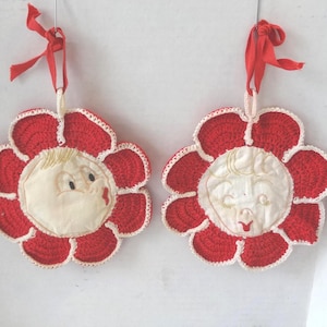 Op de afbeelding: Twee rood-witte gehaakte bloemornamenten met lachende gezichten. Elk ornament heeft een rode bloemvorm met witte rand en een crèmekleurig gezicht in het midden. Rode linten zijn bevestigd om op te hangen.