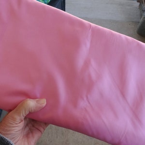 May include: A folded piece of pink fabric. The fabric has a smooth texture and a slight sheen. The colour is a soft, light pink, and the fabric appears to be a medium weight material. The fabric is being held by a hand.