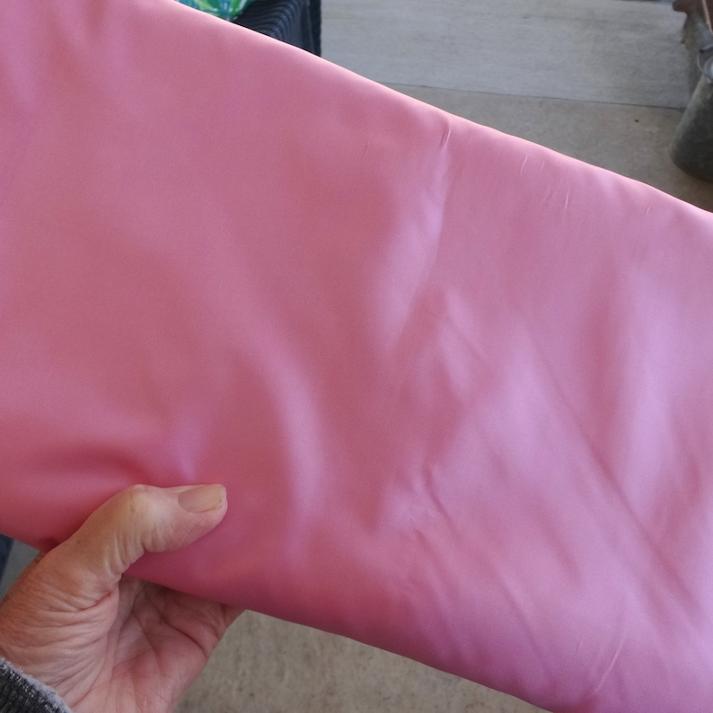 May include: A folded piece of pink fabric. The fabric has a smooth texture and a slight sheen. The color is a soft, light pink, and the fabric appears to be a medium weight material. The fabric is being held by a hand.