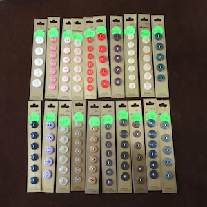 May include: A collection of sewing buttons in various colors and sizes, arranged on cardboard strips. The buttons range from white, pink, red, and purple to brown, gray, and blue. Each strip has a green sticker with a number.