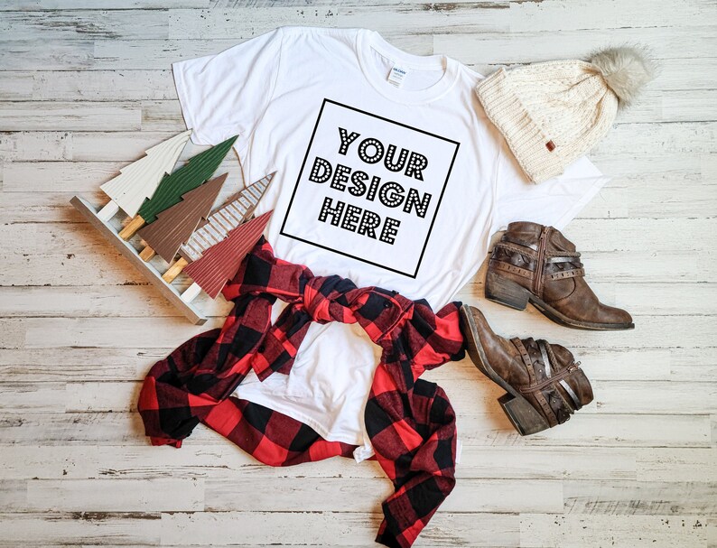 Puede incluir: Camiseta blanca con el texto "YOUR DESIGN HERE" en un cuadro negro, una camisa de cuadros rojos y negros, un gorro blanco de punto con un pomp&oacute;n marr&oacute;n y botas de cuero marr&oacute;n sobre un fondo de madera blanca.
