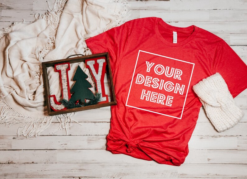 Pode incluir: Camiseta vermelha com uma caixa branca que diz "YOUR DESIGN HERE" em um fundo de madeira branca com um cobertor branco, uma placa de madeira que diz "JOY" com uma pequena &aacute;rvore de Natal verde e uma faixa de tric&ocirc; branca.