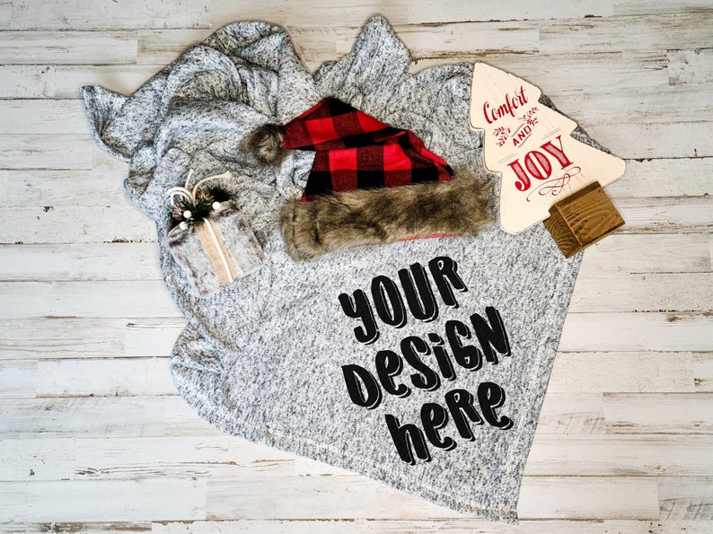 Peut inclure: Une couverture grise et blanche avec un chapeau de P&egrave;re No&euml;l en plaid rouge et noir, un petit cadeau emball&eacute; et une pancarte en bois qui dit "Confort et Joie". La couverture a les mots "Votre design ici" imprim&eacute;s dessus.