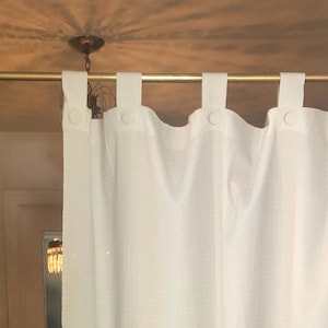 May include: A white, textured curtain with button-loop attachments hangs from a gold-colored rod. The curtain is partially drawn, revealing a glimpse of a room with a wooden table and hardwood floors. The curtain's fabric has a subtle, grid-like pattern.