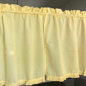 May include: Pale yellow kitchen curtains with ruffled edges and a gathered top. The curtains are hanging on a wooden rod, and the fabric appears to be lightweight. The overall style is vintage or cottagecore.