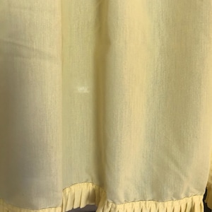May include: Pale yellow fabric with a pleated ruffle trim along the bottom edge. The fabric has a slightly textured appearance and hangs in soft folds. The ruffle trim is a lighter shade of yellow.