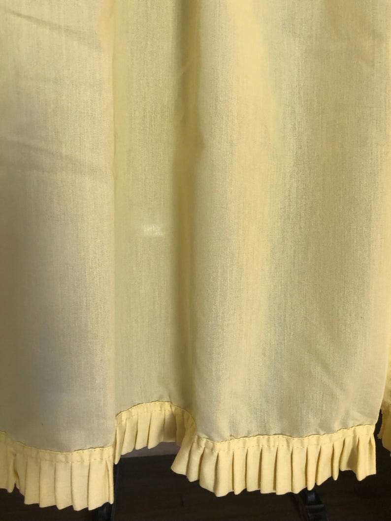 May include: Pale yellow fabric with a pleated ruffle trim along the bottom edge. The fabric has a slightly textured appearance and hangs in soft folds. The ruffle trim is a lighter shade of yellow.