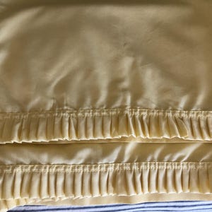 May include: Pale yellow bed sheet with pleated trim. The sheet is folded, showing the pleated detail along the bottom edge. The background is a blue and white striped pattern.