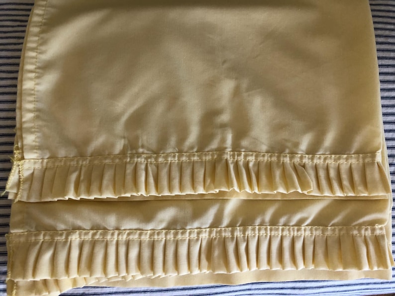 May include: Pale yellow bed sheet with pleated trim. The sheet is folded, showing the pleated detail along the bottom edge. The background is a blue and white striped pattern.