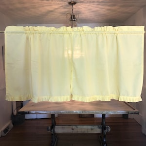 May include: Pale yellow curtains with a ruffled top and bottom edge are hanging in front of a wooden table. The curtains are gathered on a rod. The table has a light-colored top and dark, ornate legs. The room has a hardwood floor.