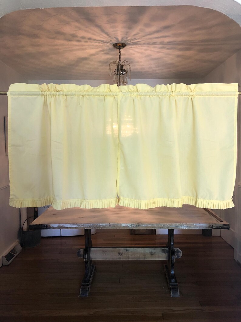 May include: Pale yellow curtains with a ruffled top and bottom edge are hanging in front of a wooden table. The curtains are gathered on a rod. The table has a light-colored top and dark, ornate legs. The room has a hardwood floor.