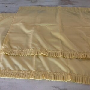 May include: Two pale yellow pillowcases with pleated ruffles along the bottom edge. The fabric has a slight sheen, and the pillowcases are neatly folded. The background is a light-colored wooden surface.