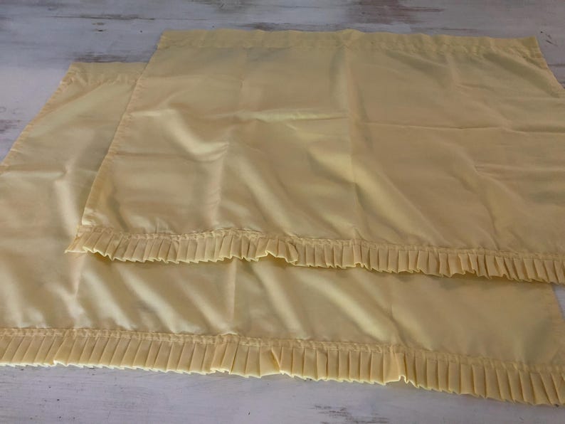 May include: Two pale yellow pillowcases with pleated ruffles along the bottom edge. The fabric has a slight sheen, and the pillowcases are neatly folded. The background is a light-colored wooden surface.