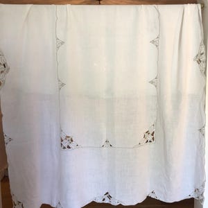May include: White linen tablecloth with a scalloped edge and decorative cut-out corners. The tablecloth features a detailed, embroidered border in a neutral color, adding a touch of elegance. The fabric is draped, showcasing its texture and design.