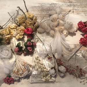 May include: Assortment of artificial flowers in various colors, including cream, pink, and red. The collection features roses, buds, and decorative elements, ideal for bridal bouquets and wedding arrangements. Some stems have attached sprigs and feathers.