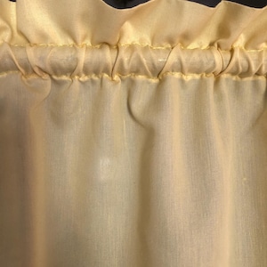 May include: Close-up of a pale yellow curtain with a ruffled top edge. The fabric is gathered and sewn with yellow thread, creating a textured, flowing appearance. The curtain is likely made of a lightweight material, suitable for filtering light.