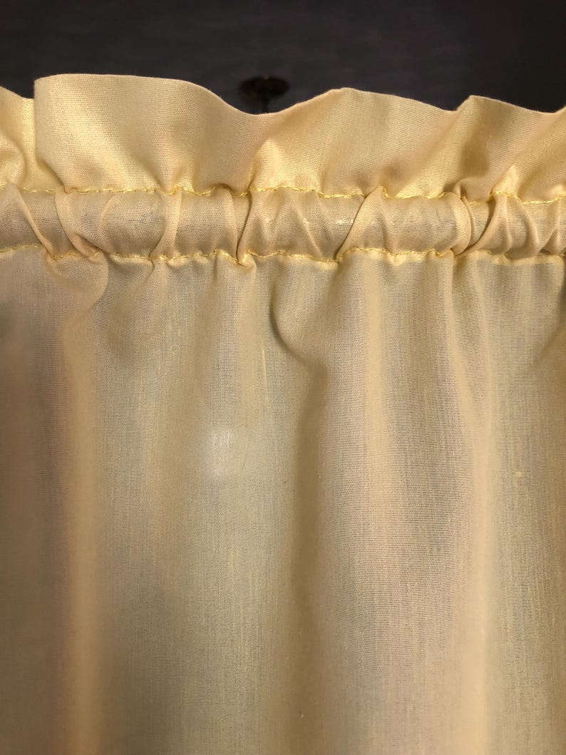 May include: Close-up of a pale yellow curtain with a ruffled top edge. The fabric is gathered and sewn with yellow thread, creating a textured, flowing appearance. The curtain is likely made of a lightweight material, suitable for filtering light.