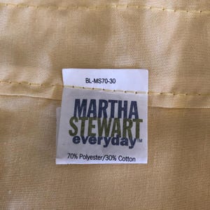 May include: Close-up of a pale yellow fabric with a tag that reads "MARTHA STEWART everyday" in green and white text. The tag also states "70% Polyester/30% Cotton". The fabric has a subtle texture and is stitched.