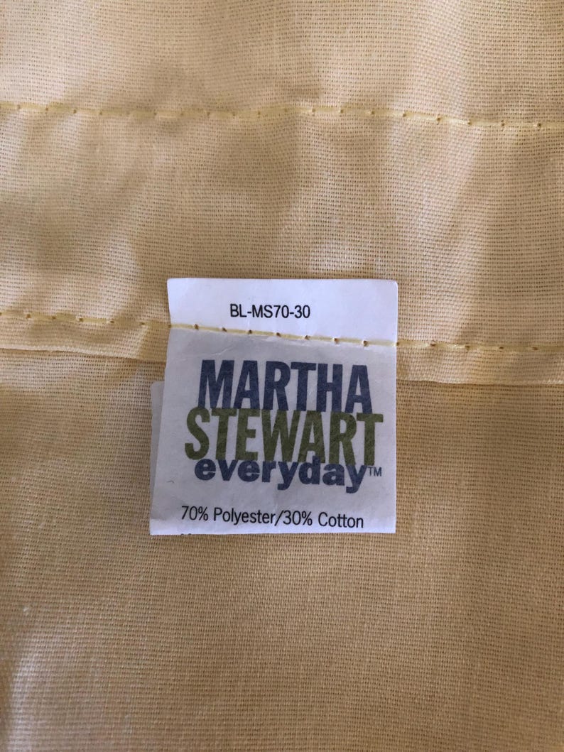 May include: Close-up of a pale yellow fabric with a tag that reads "MARTHA STEWART everyday" in green and white text. The tag also states "70% Polyester/30% Cotton". The fabric has a subtle texture and is stitched.