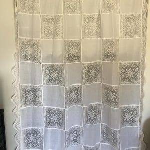 May include: A white curtain featuring a grid of alternating sheer panels and opaque panels with a floral pattern. The curtain has a scalloped edge along the sides and bottom, with a repeating star-like flower motif centered in each square panel.