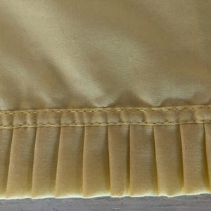 May include: Close-up of a section of pale yellow fabric with a pleated edge. The fabric has a smooth, slightly shiny texture. A line of stitching separates the smooth top from the pleated bottom.
