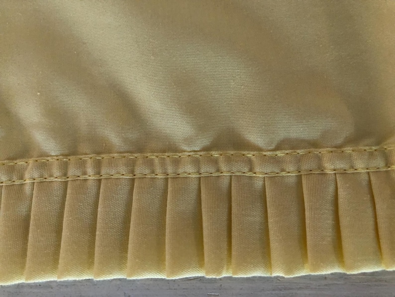 May include: Close-up of a section of pale yellow fabric with a pleated edge. The fabric has a smooth, slightly shiny texture. A line of stitching separates the smooth top from the pleated bottom.