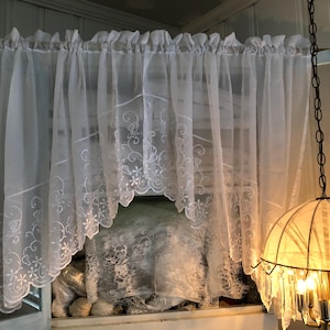 May include: A white sheer curtain valance featuring a scalloped bottom edge and intricate white floral embroidery. The fabric is lightweight and semi-transparent, with a gathered top header designed for hanging on a rod, creating a soft drape.