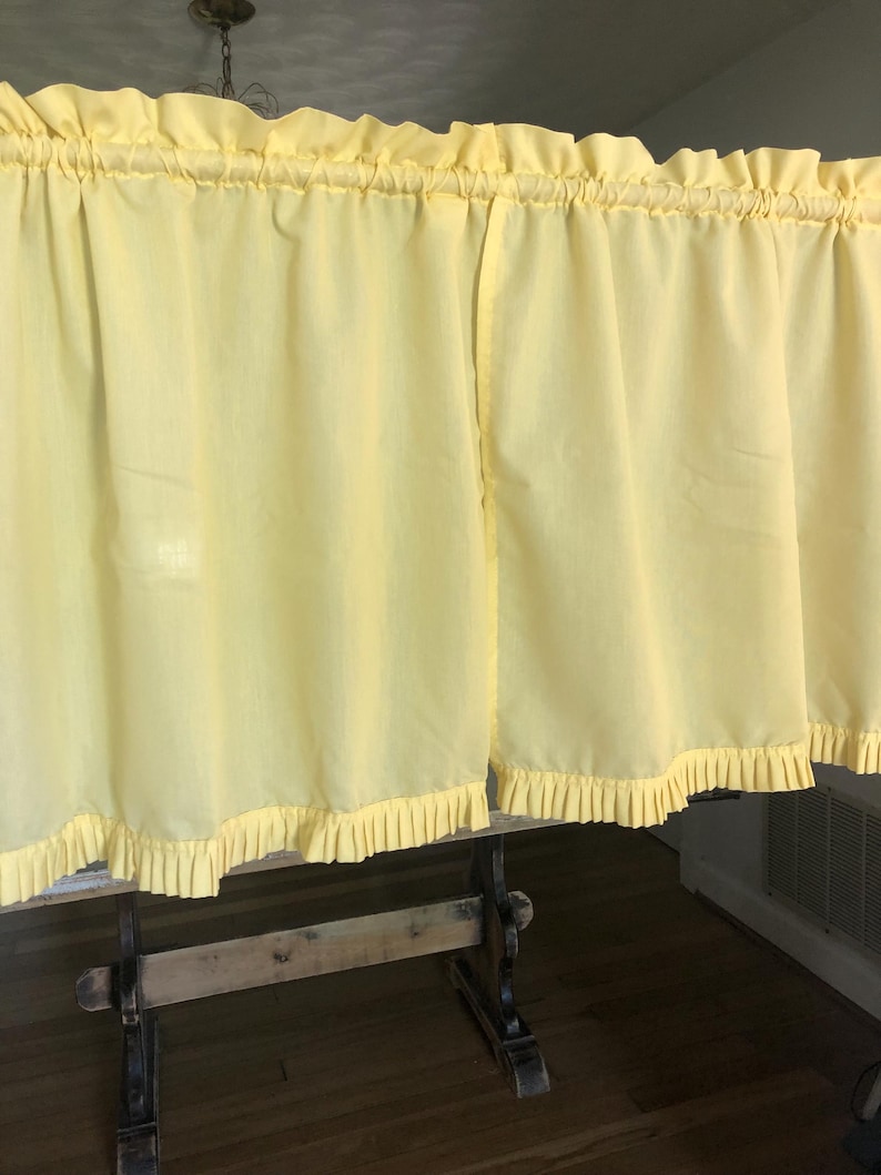 May include: Pale yellow kitchen curtains with ruffled edges and a gathered top. The curtains are hanging on a wooden rod, and the fabric appears to be lightweight. The overall style is vintage or cottagecore.
