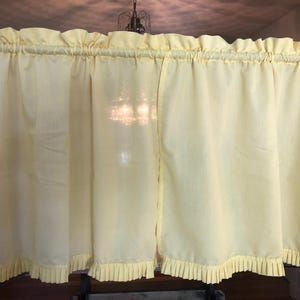 May include: Pale yellow ruffled kitchen curtains. The curtains have a gathered top and a pleated bottom edge. The fabric appears to be a lightweight material, and the curtains are hanging in front of a window.