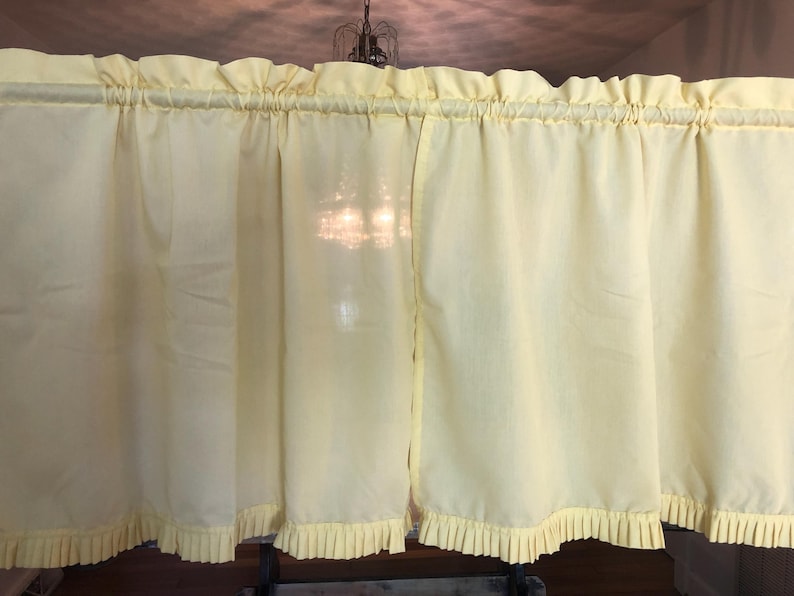 May include: Pale yellow ruffled kitchen curtains. The curtains have a gathered top and a pleated bottom edge. The fabric appears to be a lightweight material, and the curtains are hanging in front of a window.