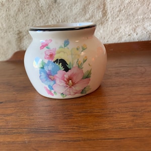 May include: A small, white ceramic vase with a rounded shape. The vase is decorated with a floral pattern featuring pink, blue, and yellow flowers. The rim is outlined with a black band. A decorative item for the home.