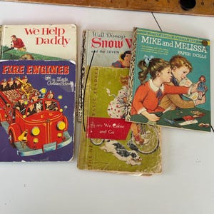 Puede incluir: Una colección de libros infantiles antiguos, incluyendo "We Help Daddy", "Fire Engines", "Snow White and the Seven" y "Mike and Melissa Paper Dolls". Los libros presentan ilustraciones coloridas y texto, centrándose en historias clásicas.