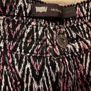 May include: Close-up of black denim pants with a pink and white zigzag pattern. The pants have a button closure and a small black tag with the Levi's logo and the words "Mid Rise Skinny".