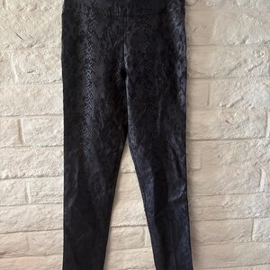 May include: Black trousers featuring a tonal snakeskin pattern throughout. These pants have a high-waisted design with a smooth waistband and a slim-fit silhouette, hanging from a clear plastic hanger against a textured white brick wall.