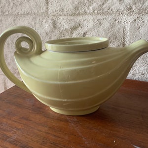 May include: A pale green ceramic teapot with a unique spiral handle and a curved spout. The teapot has a ridged design and sits on a wooden surface. The lid is a separate piece. A decorative kitchenware item.