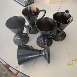 May include: Five antique silver-toned metal goblets with handles, arranged on a white surface. The goblets have a conical shape and sit on a round base. One goblet is lying on its side. The goblets appear to be vintage.