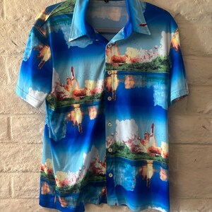 May include: A short-sleeved button-down shirt featuring a vibrant all-over print of a space shuttle launch against a bright blue sky with clouds and water reflections. The shirt has a classic collar and a label at the neck that reads KRUSH.