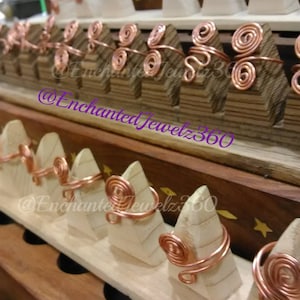 May include: Copper wire rings with spiral and swirl designs displayed on wooden stands. The rings are arranged on a tiered wooden display, showcasing the handcrafted jewelry. The rings are a warm copper color.