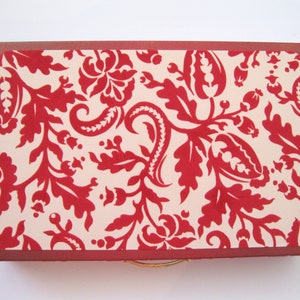 May include: A rectangular box with a red floral pattern on a cream background. The box has a dark red border and a gold-colored handle. The floral design is a repeating pattern of stylized flowers and leaves.