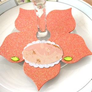 May include: A coral-colored, glittery flower-shaped coaster with a clear glass stem in the center. A small, oval-shaped card with a scalloped edge and a light pink design is in the center. Two small green gems are on the coaster.