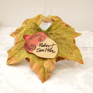 Puede incluir: Un charm para copa de vino con tem&aacute;tica oto&ntilde;al que presenta una bellota marr&oacute;n con un dise&ntilde;o de espiral rojo y una etiqueta de nombre escrita a mano que dice "Robert Smith". La bellota est&aacute; unida a una hoja de tela dorada y verde.