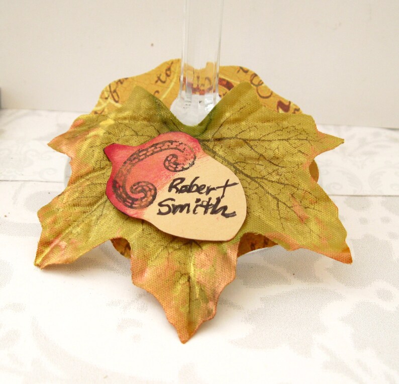 Puede incluir: Un charm para copa de vino con tem&aacute;tica oto&ntilde;al que presenta una bellota marr&oacute;n con un dise&ntilde;o de espiral rojo y una etiqueta de nombre escrita a mano que dice "Robert Smith". La bellota est&aacute; unida a una hoja de tela dorada y verde.