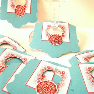 May include: Set of teal blue paper wine glass charms with a red and white snowflake design. Each charm has a circle cut out in the center for the stem of the glass.