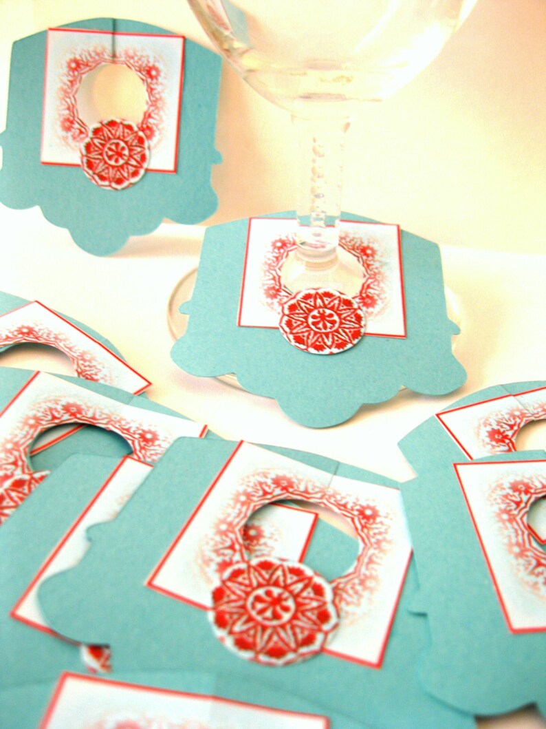 May include: Set of teal blue paper wine glass charms with a red and white snowflake design. Each charm has a circle cut out in the center for the stem of the glass.