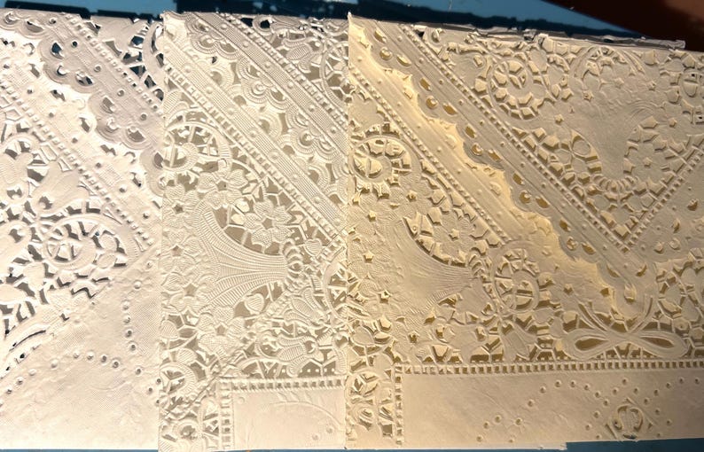 May include: Close-up of several intricately cut paper designs. The designs feature floral and geometric patterns, with delicate cut-out details. The paper has a cream color, and the designs are layered to create depth and texture.