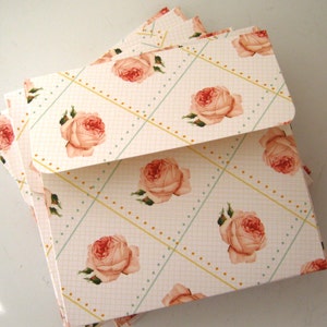 May include: A stack of white envelopes with a delicate floral pattern. The envelopes feature a repeating design of pink roses with green leaves on a white background with a subtle grid pattern and yellow and blue accents.