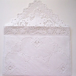 May include: A white, intricately designed envelope with a lace-like flap. The envelope features embossed floral and geometric patterns, including circles and a decorative border. The top flap has a scalloped edge, adding to its delicate appearance.