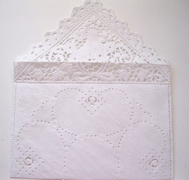 May include: A white, intricately designed envelope with a lace-like flap. The envelope features embossed floral and geometric patterns, including circles and a decorative border. The top flap has a scalloped edge, adding to its delicate appearance.