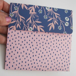 May include: A small, folded envelope with a pink dotted pattern on the lower half and a navy blue floral design on the top flap. The floral design features pink leaves and white flowers. The envelope is held by a hand.